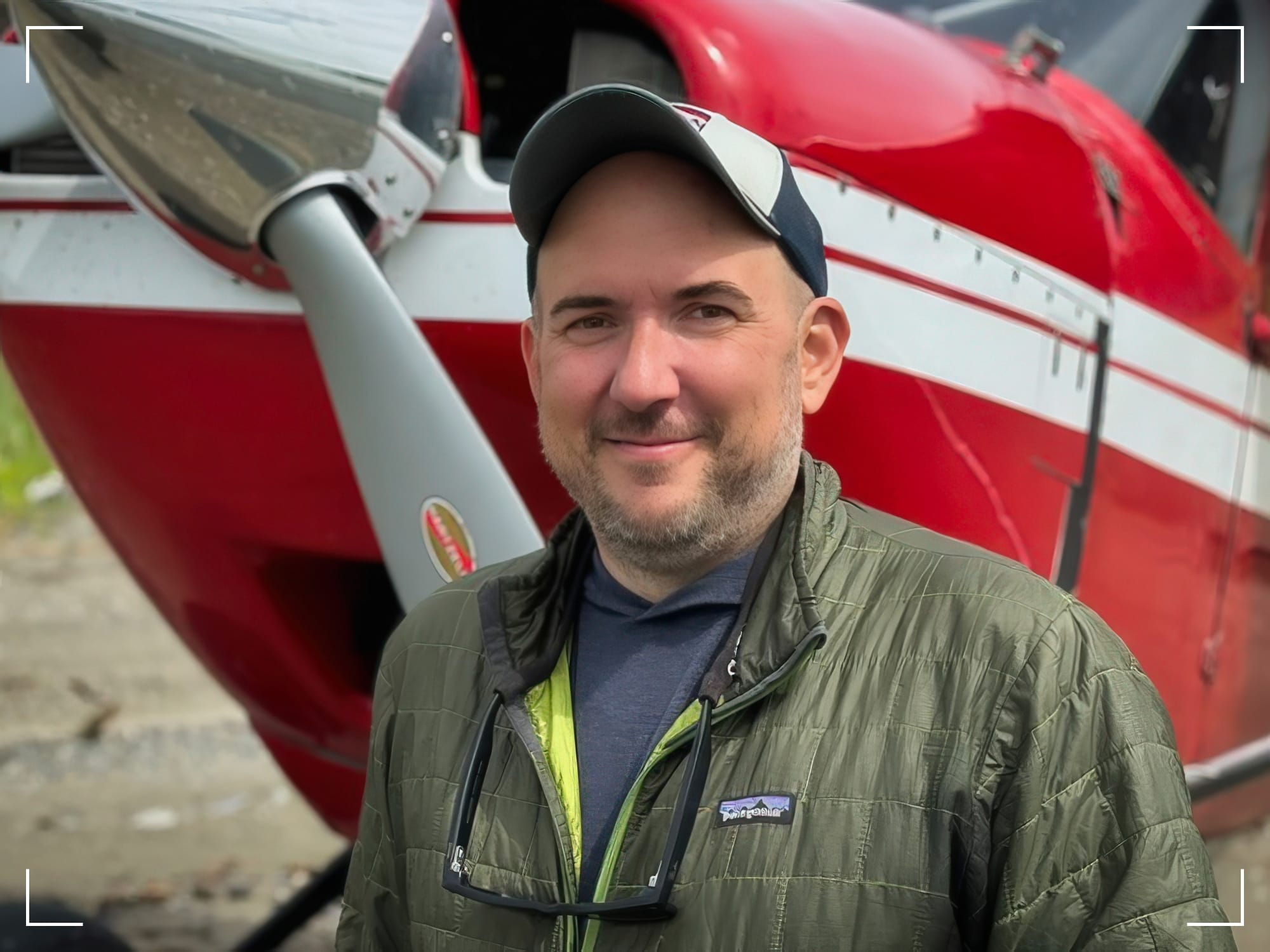 Professional photographer Zach Matthai standing in front of red and white aircraft, wearing olive jacket and baseball cap, smiling at camera