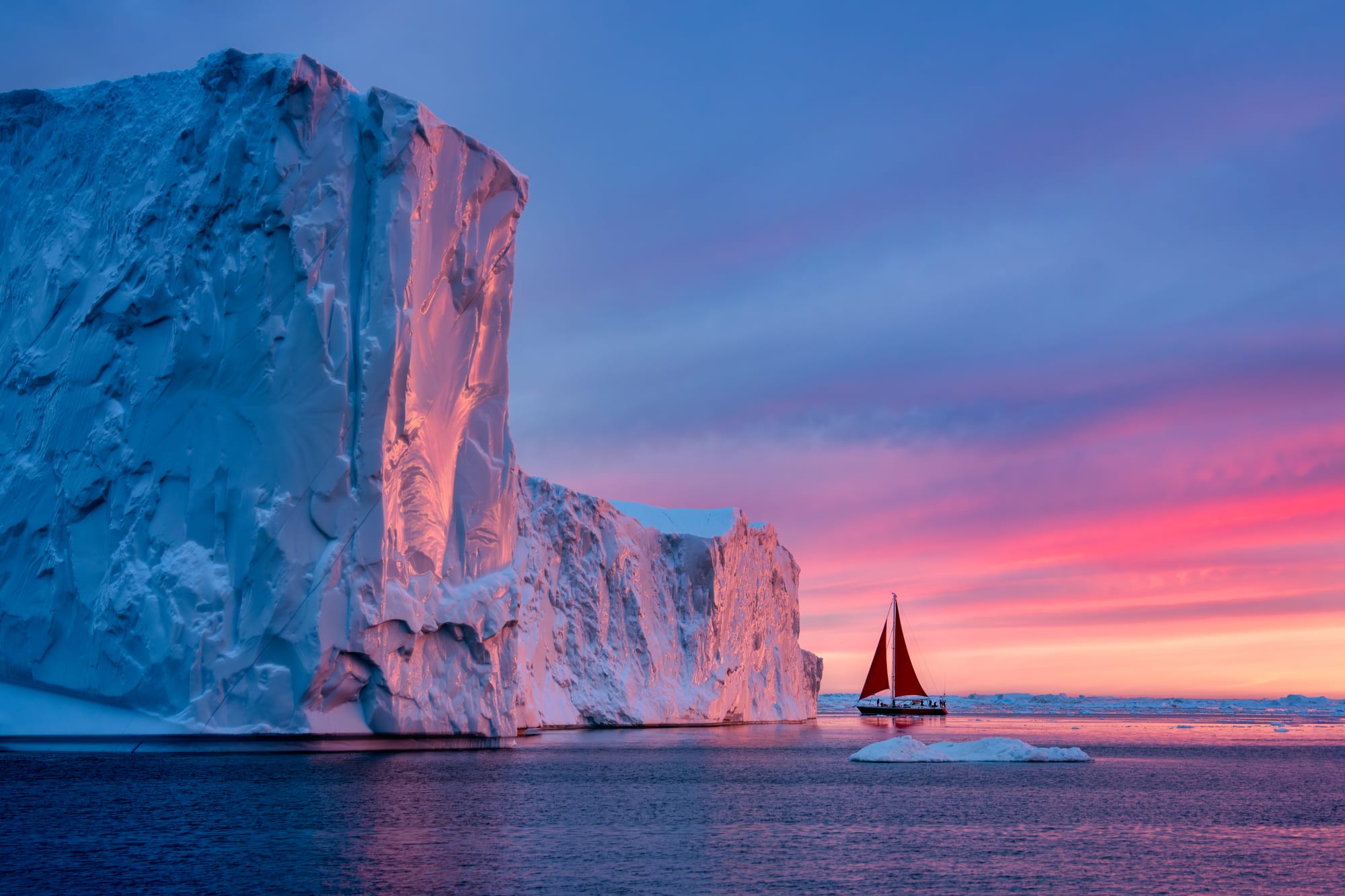 Massive iceberg in Greenland waters at sunset with sailboat showing scale